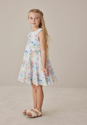 Młoda dziewczyna o długich blond włosach stoi boso, ubrana w białą sukienkę bez rękawów z kwiatowym wzorem i koronkowym kołnierzykiem, spoglądając na bok.