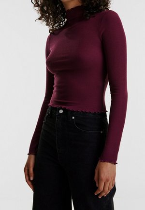 Kvinde iført en tætsiddende langærmet bourgogne ribbet top med flæsekant og højtaljede sorte jeans mod en ensfarvet baggrund.