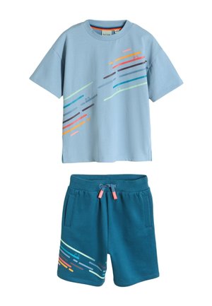 Dreng med korte dreadlocks iført en lyseblå t-shirt med grafik, blågrønne shorts med design, hvide sokker og hvide sneakers, står med hænderne i lommerne.