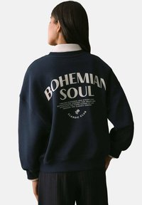 Námořnická modrá mikina s kulatým výstřihem a oversized střihem. Na zadní straně má bílý potisk "BOHEMIAN SOUL".