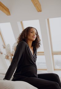 Svart pyjamas-set med vit piping, bestående av en knappad skjorta och byxor. Tyget verkar mjukt med en avslappnad passform som framhäver en gravid silhuett.