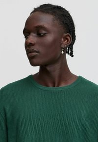 Grüner strukturierter Strickpullover mit Rundhalsausschnitt. Die Figur hat geflochtenes Haar und trägt einen silbernen Ohrring mit einem Perlenakzent.