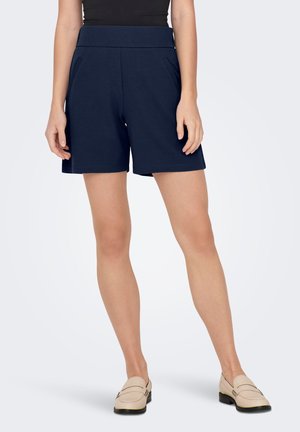 Vrouw draagt marineblauwe op maat gemaakte shorts, zwarte top en beige loafers, staand tegen een effen lichte achtergrond.