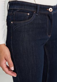 Jeans de mezclilla azul oscuro con herrajes de cobre, que cuentan con un diseño clásico de cinco bolsillos y costuras en contraste a lo largo de las costuras y los bolsillos.