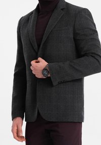 Ciemnoszara, teksturowana marynarka o dopasowanym kroju, z klapą z wycięciem, jedną kieszenią na piersi i guzikami. Nosi się ją na dopasowany golf w tym samym kolorze.