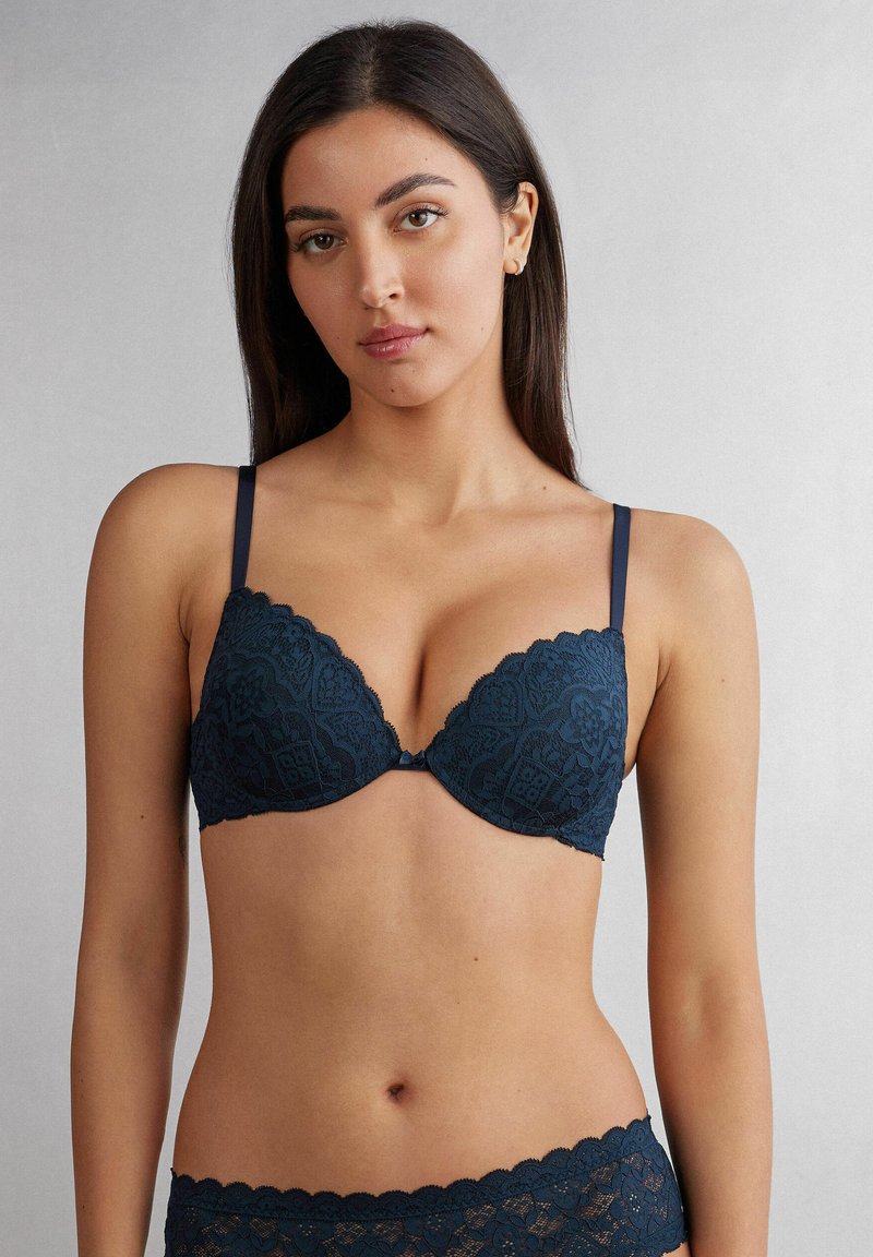 Marinblå spets-bh med vadderade, triangulära kupor, scalloperade kanter och justerbara axelband, prydd med ett blommönster och en central rosett.