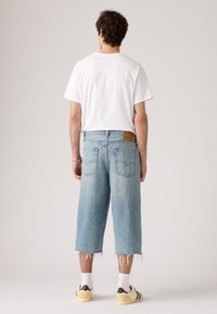 Shorts de mezclilla azul claro con un corte holgado, dobladillo sin rematar y logo de parche en la parte posterior. Combinados con una camiseta blanca y zapatillas crema.