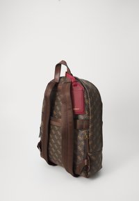 Sac à dos brun à motifs avec des sangles en tissu brun, poches zippées et une étiquette en métal rouge. Comprend des détails de logo et une surface texturée.