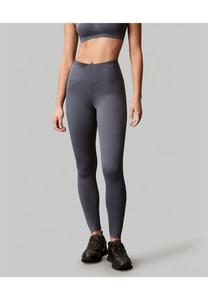 Kobieta w ciemnoszarych legginsach z wysokim stanem, dopasowanym sportowym biustonoszu i czarnych butach sportowych na jednolitym szarym tle.