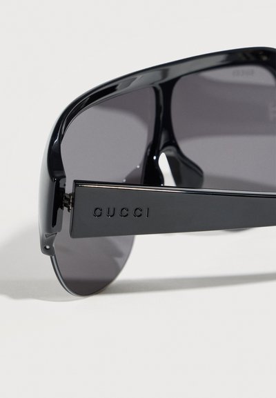 Schwarze Gucci-Sonnenbrille mit breiten Bügeln und dunkel getönten Gläsern, in Nahaufnahme vor weißem Hintergrund gezeigt.