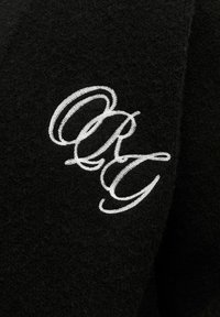 Schwarzer Wollstoff mit einem auffälligen silbernen gestickten "ORG"-Logo. Der Stoff hat eine weiche, leicht strukturierte Oberfläche.