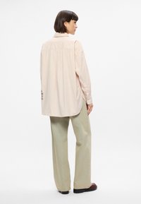 Ανοιχτό ροζ oversized πουκάμισο με γιακά, μακριά μανίκια και πλισέ πίσω. Διαθέτει κεντημένες λεπτομέρειες κοντά στο στρίφωμα. Συνδυάζεται με ανοιχτό πράσινο παντελόνι.