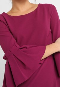 Bordeauxklänning med båtkrage och utsvängda ärmar med veckade detaljer. Tillverkad av ett mjukt och slätt tyg.