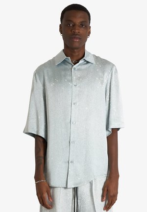 Uomo con camicia azzurro chiaro a maniche corte abbottonata e pantaloni abbinati azzurro chiaro su sfondo uniforme.