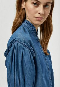 Denim srajca z valovitim ovratnikom in napihnjenimi rokavi v srednje modri barvi. Ima sprednje gumbe in gladko teksturo.