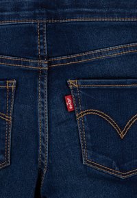 Jeans in denim blu scuro con cuciture dorate, due tasche posteriori e un'etichetta rossa del marchio sulla tasca destra.