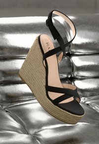 Black wedge sandal with crisscross straps, woven jute sole, and smooth leather texture. Accent of adjustable ankle strap and open toe design.