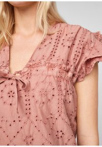 Blusa ricamata color rosa con scollatura a V, caratterizzata da motivi floreali e dettagli in pizzo. Maniche corti con volant e un fiocco decorativo.