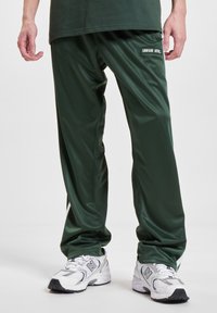 Homme portant un pantalon de sport vert foncé avec un écusson indiquant "UNFAIR ATHL" et des baskets blanches New Balance.