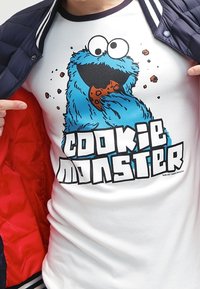 Osoba ubrana w białą koszulkę z rysunkowym Cookie Monsterem trzymającym ciastko oraz napisem "COOKIE MONSTER" poniżej.