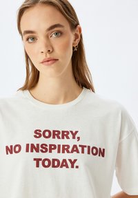 Hvid bomulds t-shirt med fed burgunder tekst, der står: "BEKLAGER, INGEN INSPIRATION I DAG." Rund hals og afslappet pasform.