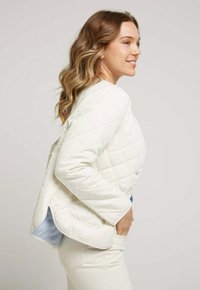Veste blanche matelassée avec un col rond, des poches latérales et une doublure rayée bleue visible à l'ourlet. Texture lisse et coupe ajustée.
