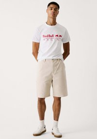 Witte T-shirt met rood "Red Bull Racing" logo, beige shorts, witte schoenen met zwarte accenten en witte sokken. Eenvoudige, casual outfit.