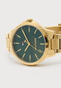 Ρολόι σε χρυσό τόνο με πράσινο καντράν, που διαθέτει χρυσούς δείκτες ωρών, λογότυπο στις 12 η ώρα και μεταλλική αλυσίδα. Χωρίς επιπλέον σχέδια ή διακοσμητικά στοιχεία.