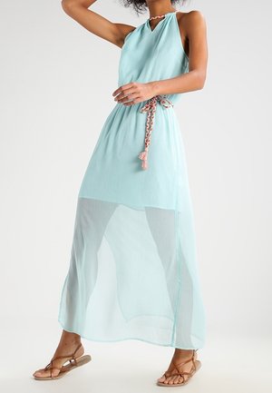 Kobieta w bezrękawnej, jasnoniebieskiej sukience maxi na szyję z przezroczystą nakładką na spódnicę i plecionym paskiem, w parze z brązowymi sandałami na paski.