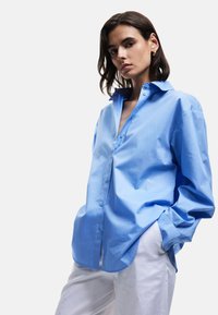 Camicia azzurro chiaro con bottoni, dal fit rilassato, con colletto a punta, maniche lunghe e una texture liscia e leggermente lucida.