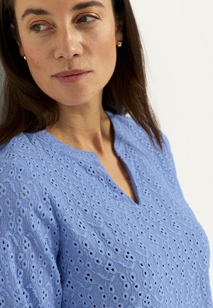 Kvinde med langt brunt hår iført små guldøreringe og en himmelblå top af øjlet stof med en hakket halsudskæring, der ser til siden.