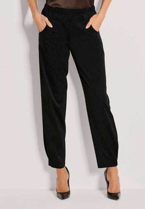 Femme portant un pantalon noir taille haute avec une ceinture élastique et des poches, associé à des talons noirs pointus, debout contre un mur clair.