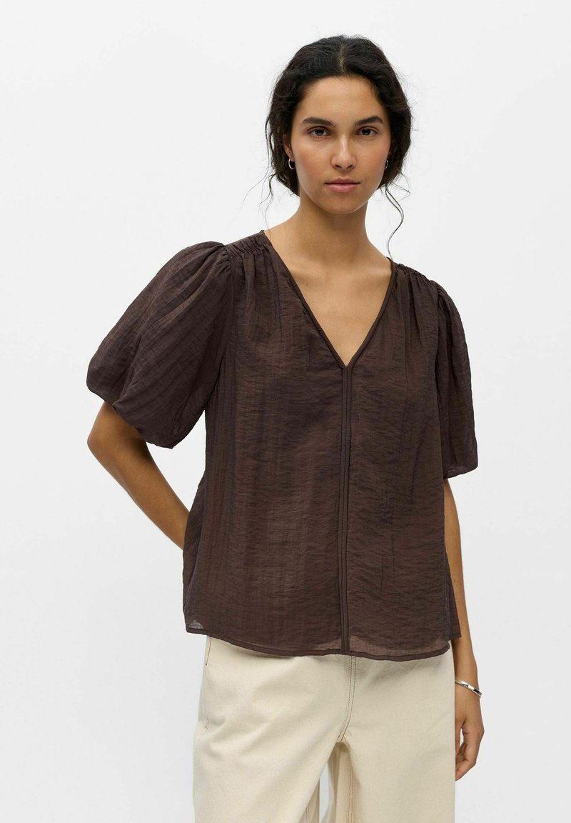 Mujer con cabello rizado oscuro que lleva una blusa marrón transparente con escote en V y mangas abullonadas, y pantalones beige claro, de pie frente a un fondo liso.