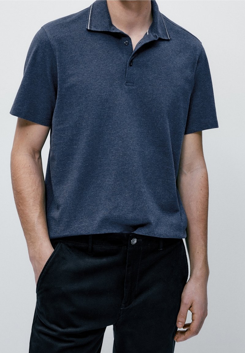 Polo à manches courtes en coton bleu foncé avec une finition texturée ; doté d'un col contrastant et d'une patte de boutonnage à trois boutons. Associé à un pantalon noir.