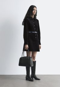 Schwarzes Cordhemdkleid mit Kragen und Knopfverschluss, tailliert mit einem hellblauen Gürtel. Kombiniert mit knielangen schwarzen Stiefeln und einer Handtasche.