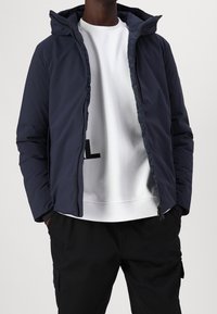 Marineblå vindjakke med hætte, der har en fuld lynlås og elastiske manchetter, lagd ovenpå en hvid sweatshirt med en fed sort skrifttype.