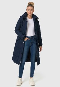 Navy pufferjas met een capuchon, ritssluiting aan de voorkant en knoopsluiting. Gelaagd ontwerp met gestikte textuur en lange lengte, gecombineerd met blauwe jeans.