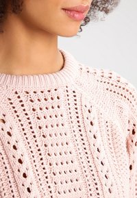 Pull en maille rose clair avec un motif ajouré, présentant un col côtelé et un design texturé, mettant en valeur un tissu doux et respirant.