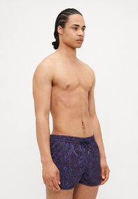 Shorts de bain violets avec un motif abstrait sombre contrastant, taille élastique et cordon de serrage. Le matériau semble léger et flexible.