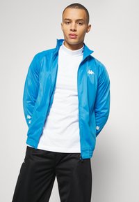 Modrá športová bunda s zipsom a hladkou textúrou, s bielym logom značky na hrudi a rukáve, v kombinácii s čiernymi nohavicami.