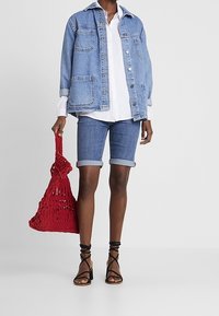 Mujer vistiendo shorts de mezclilla remangados, chaqueta de mezclilla oversize, camisa blanca, tacones negros de tiras, sosteniendo un bolso tote tejido rojo.