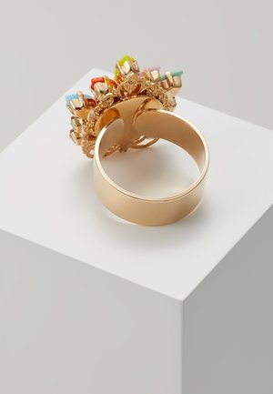 Bague en or avec un large anneau présentant un groupe décoratif de pierres précieuses rectangulaires multicolores sur le dessus, exposée sur un socle blanc.