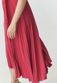 Abito plissè rosso con un design fluido. Il tessuto appare morbido con una superficie liscia. Un dettaglio in nastro è visibile in vita.