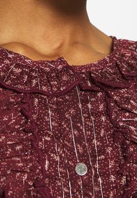 Top bordeaux con scollatura arricciata, caratterizzato da un motivo floreale e strisce argentate, fissato con un bottone metallico al centro davanti.