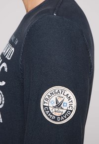 Tengerészeti kék, hosszú ujjú ing, texturált anyagból, fehér logónyomattal és a bal ujján egy kör alakú folttal, amely tengeri dizájnelemeket tartalmaz.