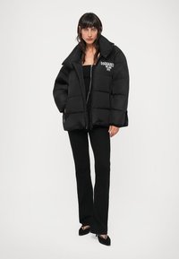 Femme portant une doudoune noire avec le texte "DSQUARED MILANO 1995", un haut noir, un pantalon noir et des talons aiguilles noirs, se tenant devant un fond blanc.