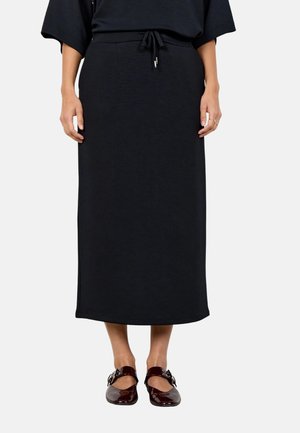 Woman wearing a black midi skirt with a tie waist and dark red patent leather Mary Jane shoes with buckles.