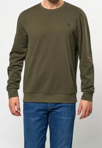 Olivi vihreä pitkähihaisessa crew neck -hupparissa on pehmeä kangas, jossa on ribbineuloksiset hihansuut ja helma, sekä pieni sininen logo vasemmassa rinnassa.