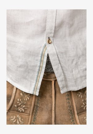 Weißes Leinenhemd mit abgerundetem Saum, ausgestattet mit einer Knopfleiste und einem mehrfarbigen gestreiften Akzent. Darüber auf einem sandfarbenen Stoff mit floralen Stickereien.