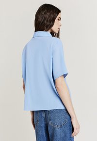 Donna con lunghi capelli scuri, che indossa una maglietta a maniche corte di un leggero blu texture e jeans di denim blu, rivolte di lato a uno sfondo neutro.